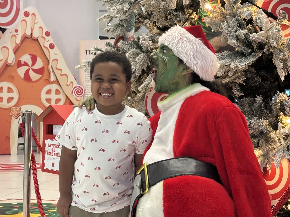 Enfant souriant avec le Grinch devant une maison en pain d'épices lors d'une animation de Noël à La Réunion